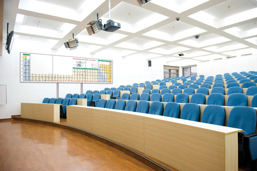 Großer Hörsaal mit blauen Sitzreihen und blume Periodensystem-Wandklapptafel an der Stirnwand